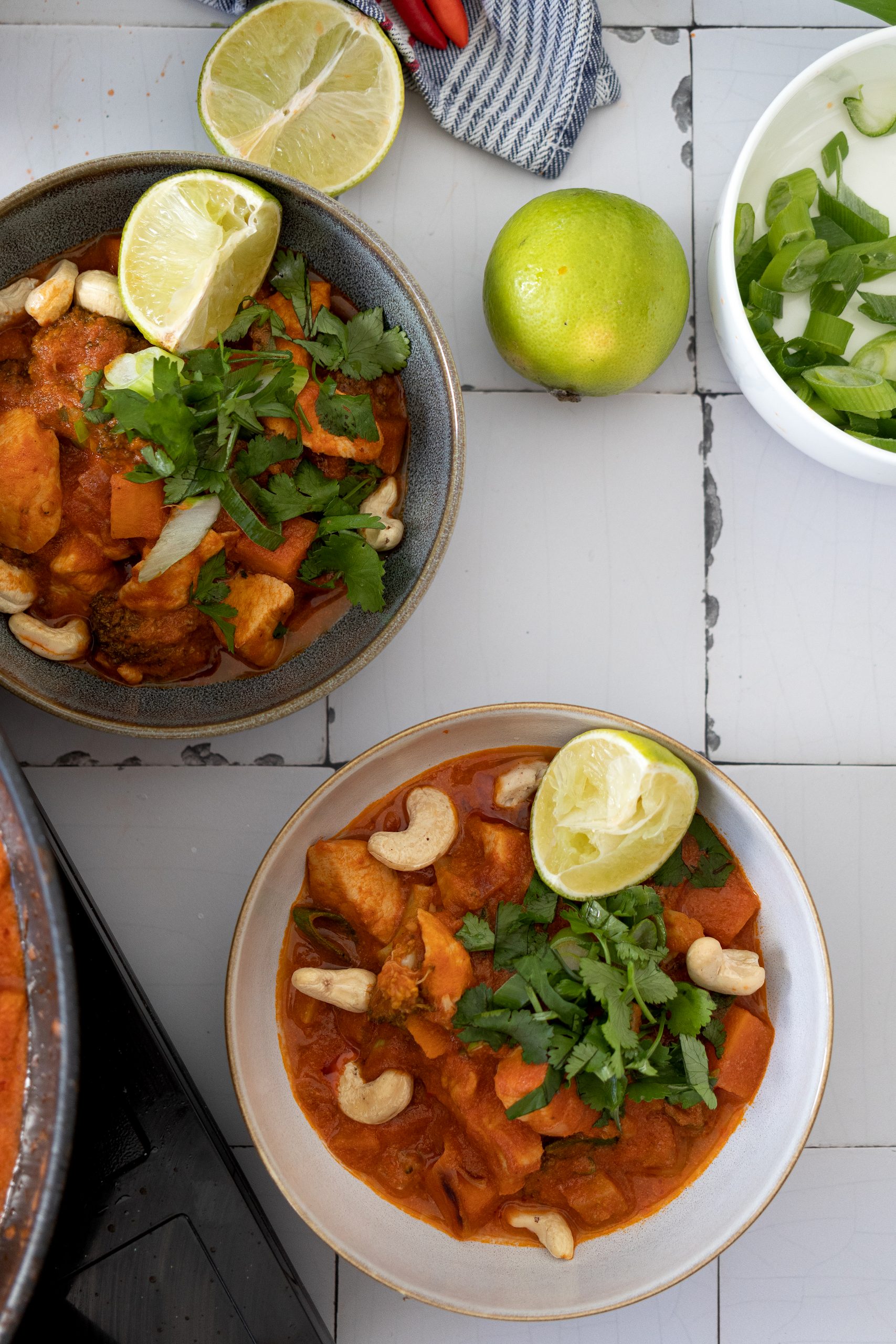 One Pot Süßkartoffel Hähnchen Zwei Schalen mit Curry aus Gemüse, Cashewnüssen und Limettenscheiben, daneben eine ganze Limette und Frühlingszwiebeln.