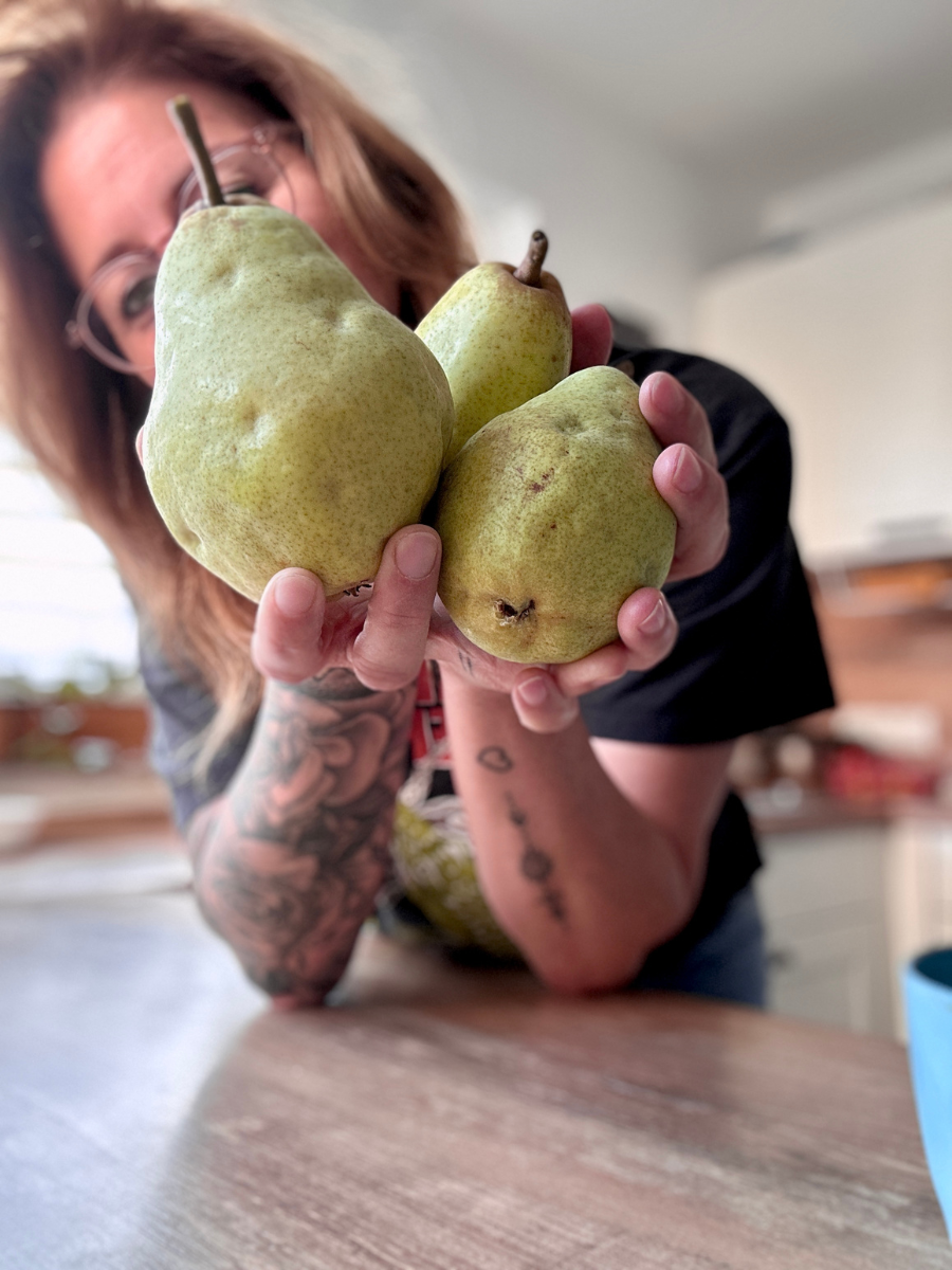 Birnenauflauf-02 Person mit Brille und Tattoos zeigt drei grüne Birnen in einer Küche.