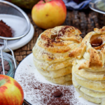 Blätterteig-Äpfel mit Zimt, dekoriert auf einem Teller, daneben Kakaopulver und Schokotropfen in einem Sieb.