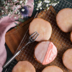 Runde Plätzchen mit Zuckerguss auf einem Gitterrost, eines teils mit rosa Glasur, neben einer Gabel und Blumen.