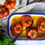 Gefüllte Tomaten in einer Ofenform auf Fliesen, umgeben von frischen Kräutern und Frühlingszwiebeln.
