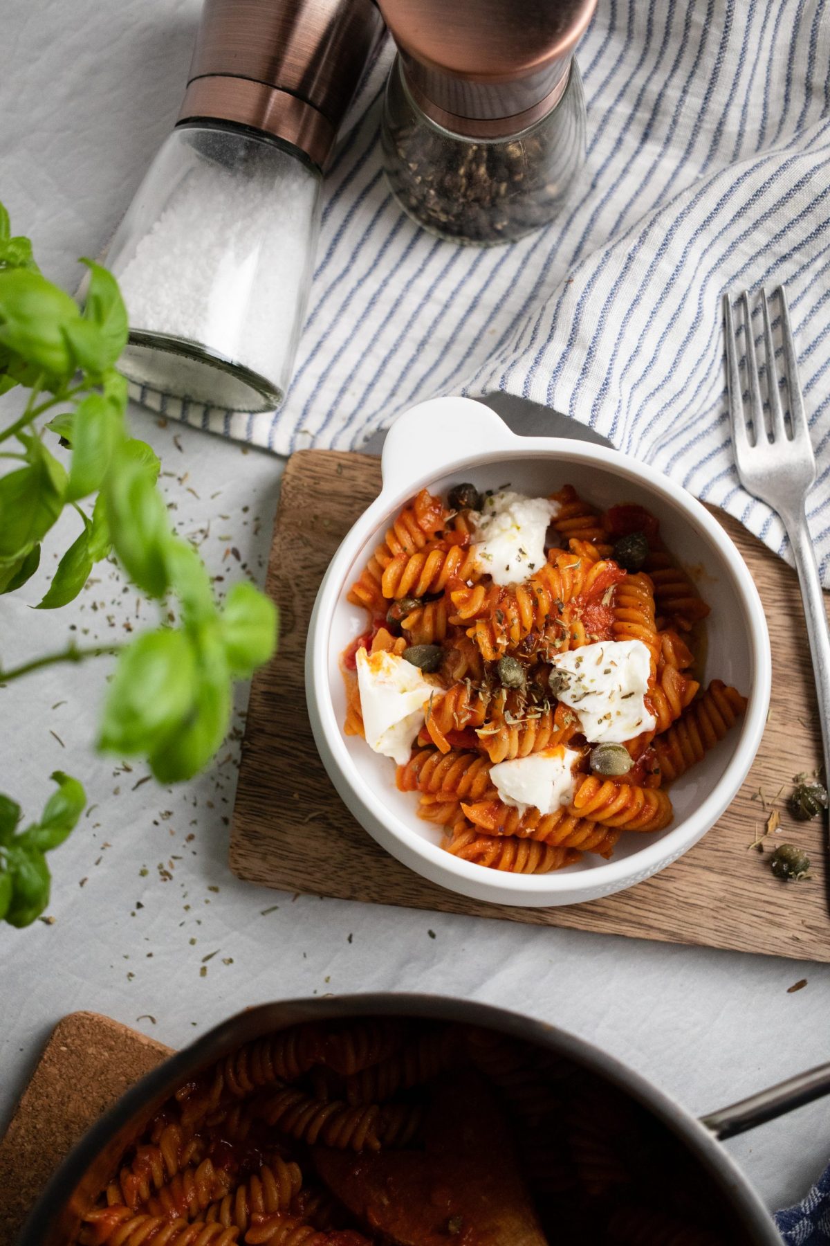 One Pot Pasta Schüssel mit Fusilli in Tomatensauce, garniert mit Käse und Kapern, neben Basilikum und Gewürzmühlen auf gestreiftem Tuch.