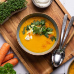 Schale mit Karottensuppe, dekoriert mit Kräutern und Sahne, auf Holzbrett mit Kresse, Karotten und Löffeln.