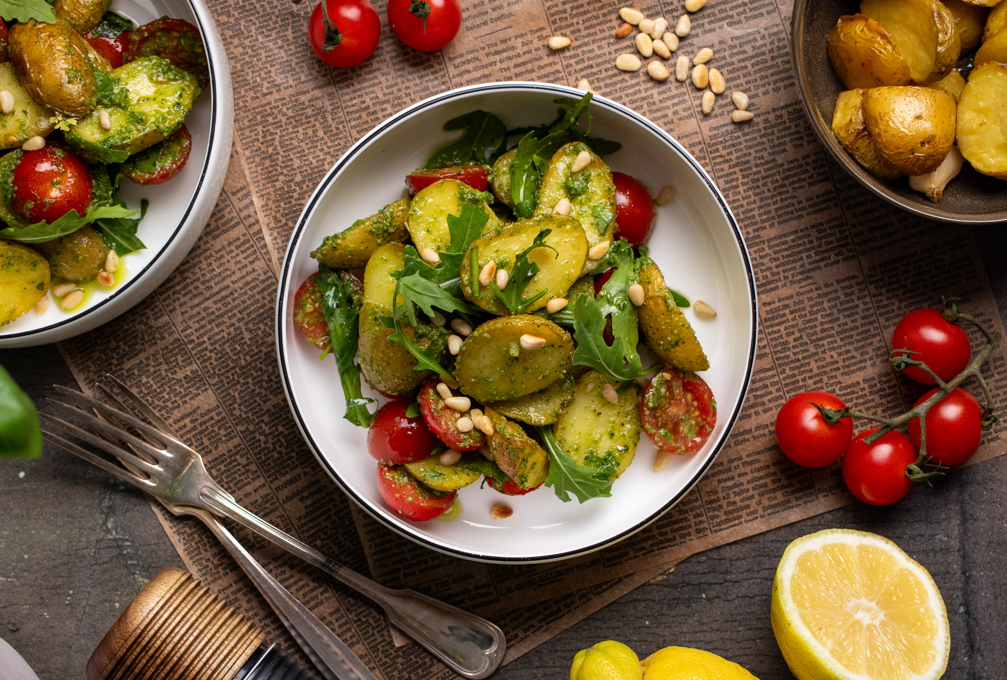 Backkartoffelsalat_01 Schüssel mit Kartoffelsalat, Tomaten, Rucola und Pinienkernen auf Zeitungspapier, daneben Cherrytomaten und halbe Zitrone.