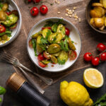 Schüsseln mit Kartoffel-Rucola-Salat und Kirschtomaten, geröstete Kartoffeln, Pinienkerne und Zitronen auf dunklem Untergrund.