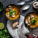 Zwei Schalen gelbe Suppe mit Pilzen, Frühlingszwiebeln und Koriander auf Stofftuch, umgeben von Limetten und Champignons.