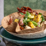 Tortilla-Schale mit Avocado, Mais, Speck und Koriander auf einem grünen Teller, vor grünem Hintergrund.