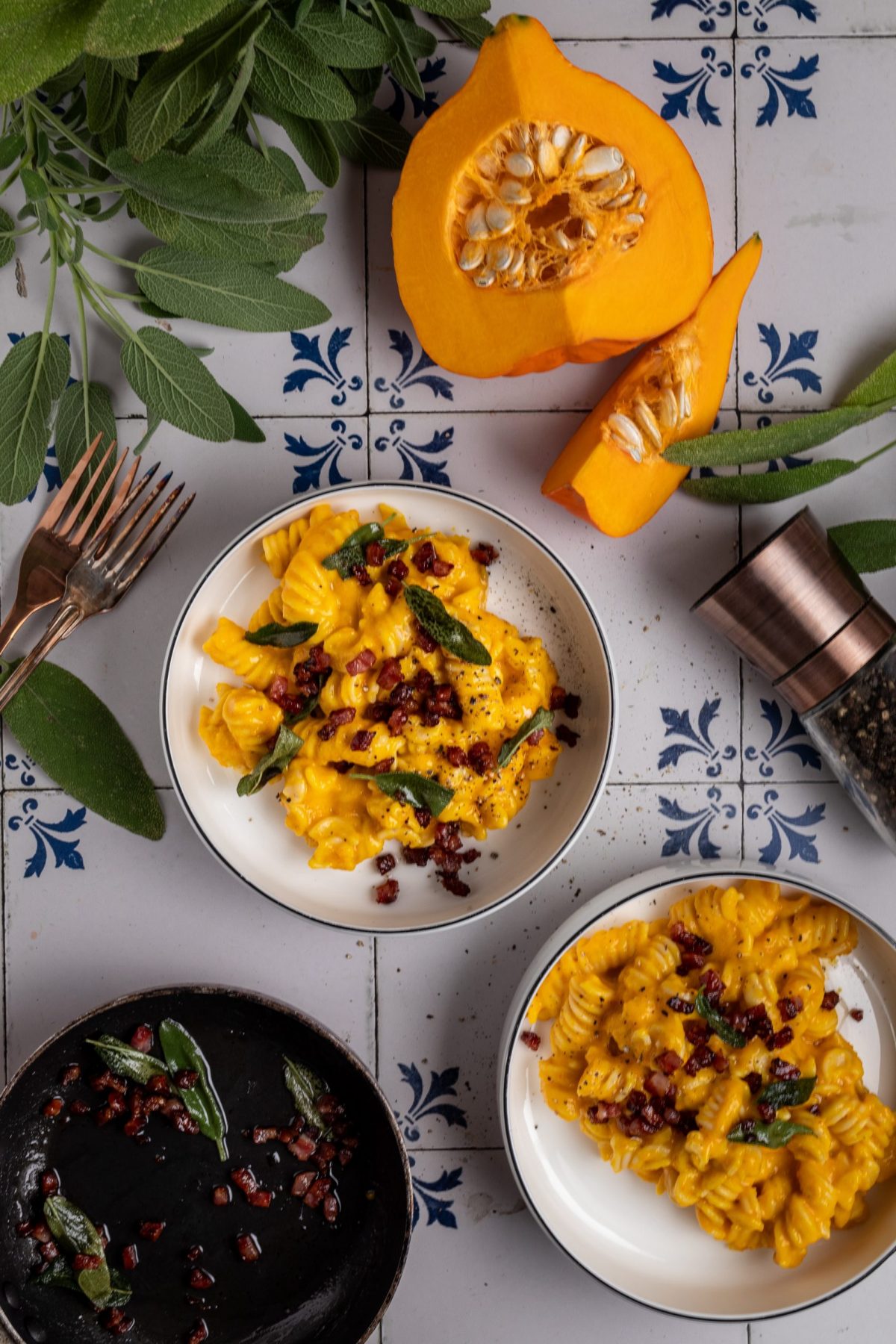 Gekochte Pasta in Kürbissoße mit Speck und Salbei, auf Fliesentisch, umgeben von Kürbisstücken und Kräutern.