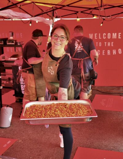 Frau mit Brille und Schürze präsentiert glücklich ein Tablett voller Nudeln in rotem Zelt. Zwei weitere Personen im Hintergrund.