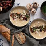 Zwei Schalen mit cremiger Suppe, Pilzen, Croutons und Schnittlauch, daneben Brotlaib, Champignons und grüne Kräuter.