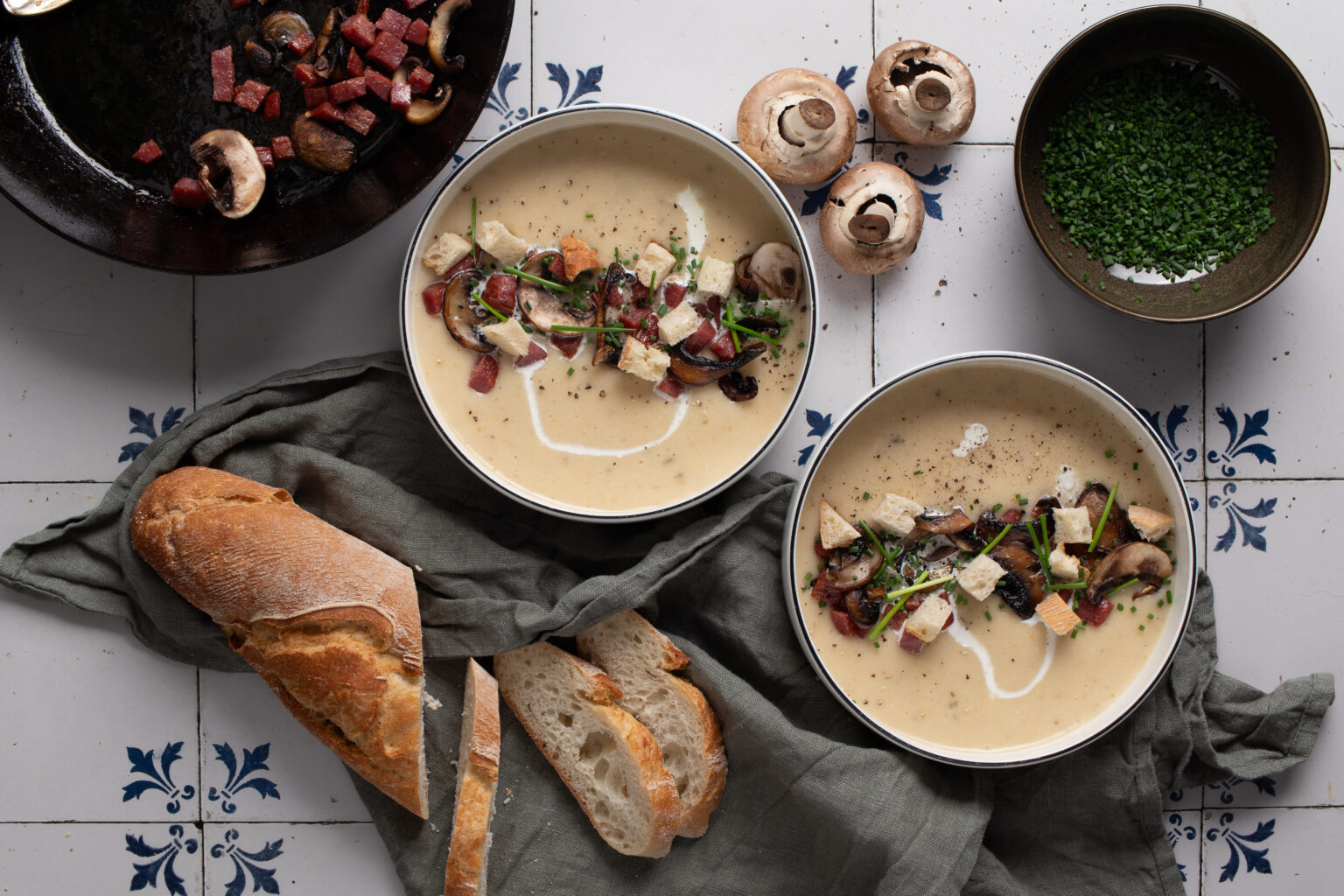 Zwei Schalen mit cremiger Suppe, Pilzen, Croutons und Schnittlauch, daneben Brotlaib, Champignons und grüne Kräuter.