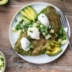 Grünes Omelett mit Avocado, Gurkenwürfeln und Kräutern, garniert mit weißen Soßenklecksen auf einem weißen Teller.