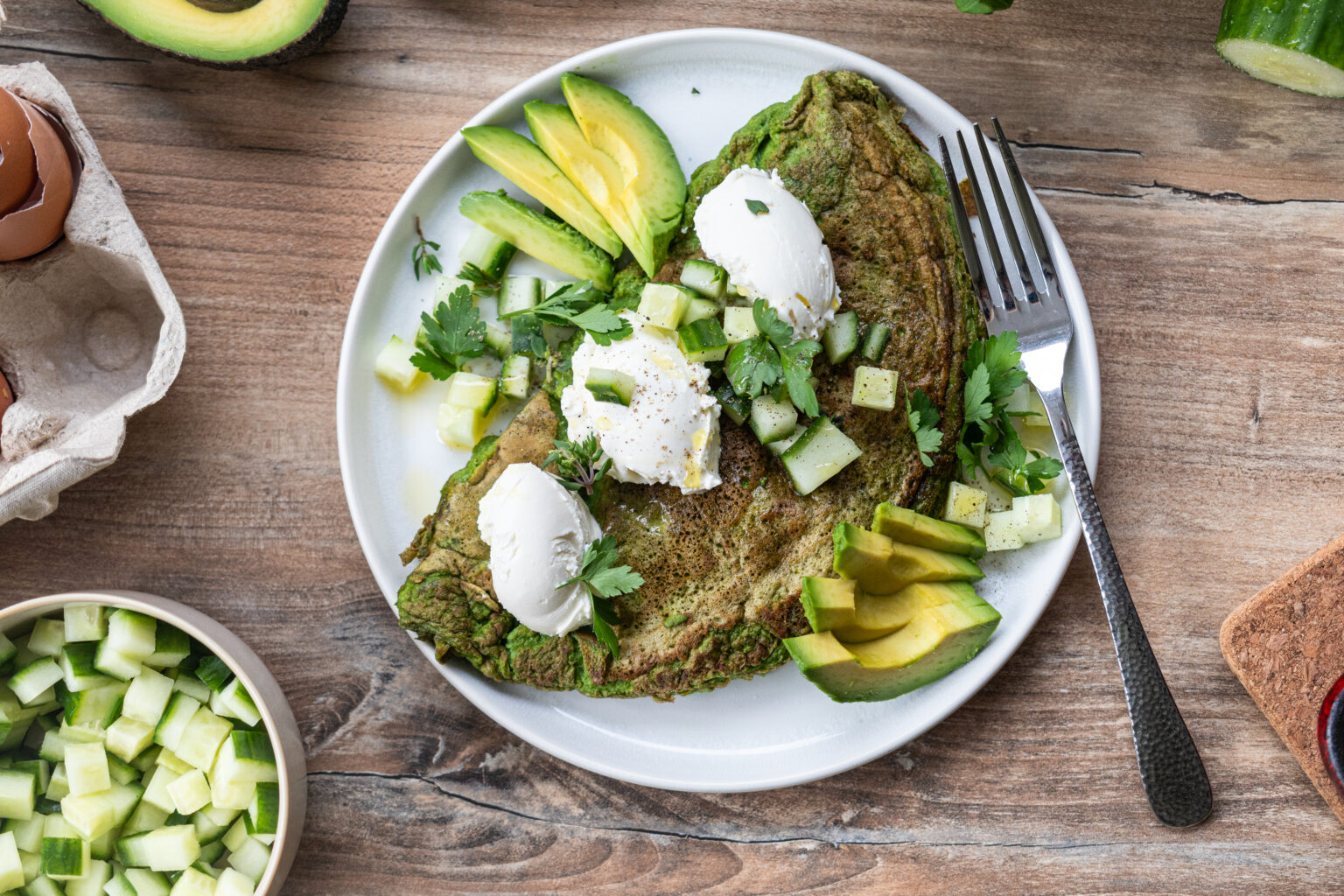 Grünes Omelett mit Avocado, Gurkenwürfeln und Kräutern, garniert mit weißen Soßenklecksen auf einem weißen Teller.