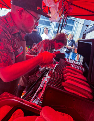 Mann grillt Würstchen unter rotem Zelt; weitere Person und Menschen im Hintergrund.