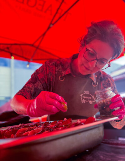 Person mit Brille und Handschuhen würzt Essen in einer großen Metallschale unter einem roten Zelt.