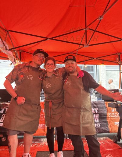 Drei Personen in Schürzen posieren fröhlich unter einem roten Pavillon.