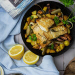 Gebratener Fisch mit Kapern und Kartoffeln in einer Pfanne, daneben Zitronenhälften und ein Glas mit eingelegten Gurken.