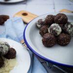 Schokoladen-Energie-Bällchen auf einem Teller, teils in Mandelmehl gewälzt, mit Datteln im Hintergrund.