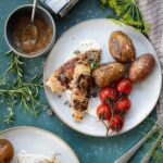 Gegrillter Fisch mit Tomaten und Kartoffeln auf Teller, daneben Kräuter und eine Schüssel mit Sauce, auf dunkelblauem Untergrund.