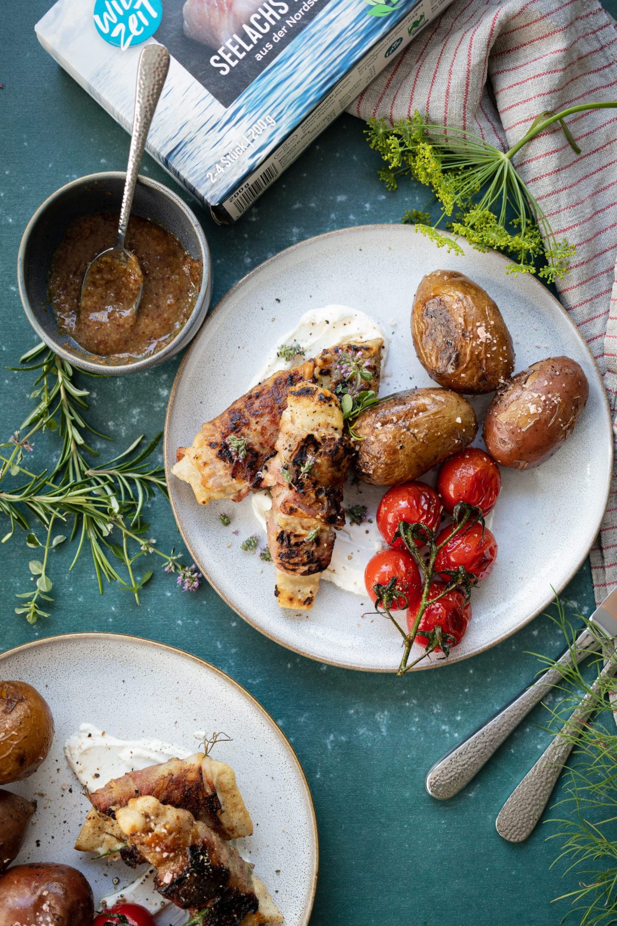 Gegrillter Fisch mit Tomaten und Kartoffeln auf Teller, daneben Kräuter und eine Schüssel mit Sauce, auf dunkelblauem Untergrund.
