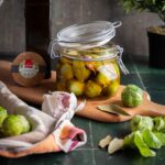 Ein Glas mit eingelegtem Rosenkohl, Knoblauch und Lorbeerblättern, neben einer Ölflasche auf einem Holzbrett.