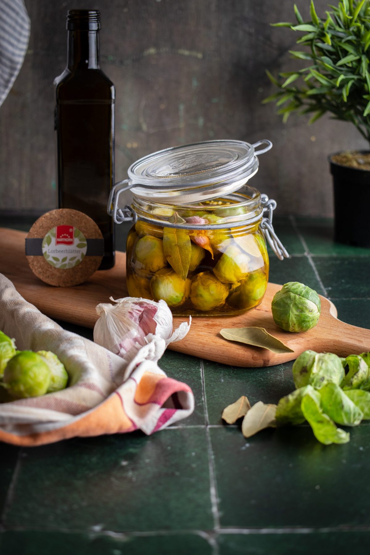 Ein Glas mit eingelegtem Rosenkohl, Knoblauch und Lorbeerblättern, neben einer Ölflasche auf einem Holzbrett.