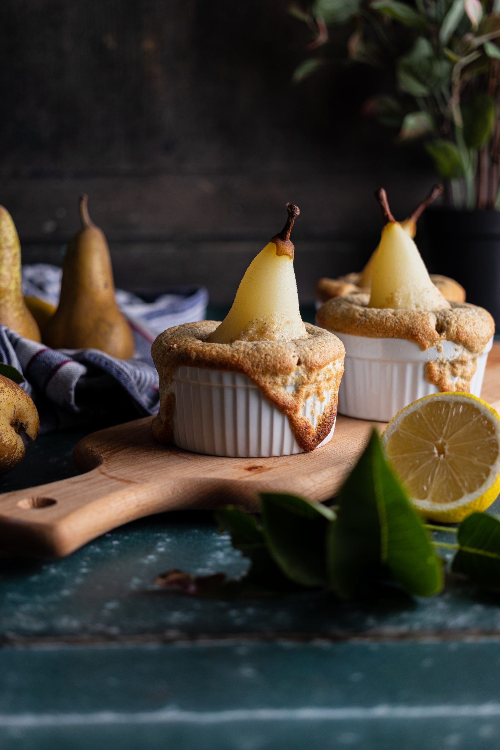 Zwei Birnen-Küchlein in Förmchen auf Holzbrett, umgeben von Birnen und Zitronenhälfte, rustikaler Hintergrund.