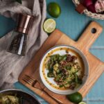 Salat mit Gurken und Gewürzen in einer Schale auf Holzbrett, umgeben von Limetten, Pfeffermühle und Gemüse auf blauem Tisch.