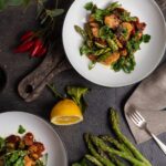 Zwei Teller mit Hühnchen, Spargel und Gemüse, umgeben von Zitronen, Chili und frischen Kräutern auf dunklem Holztisch.