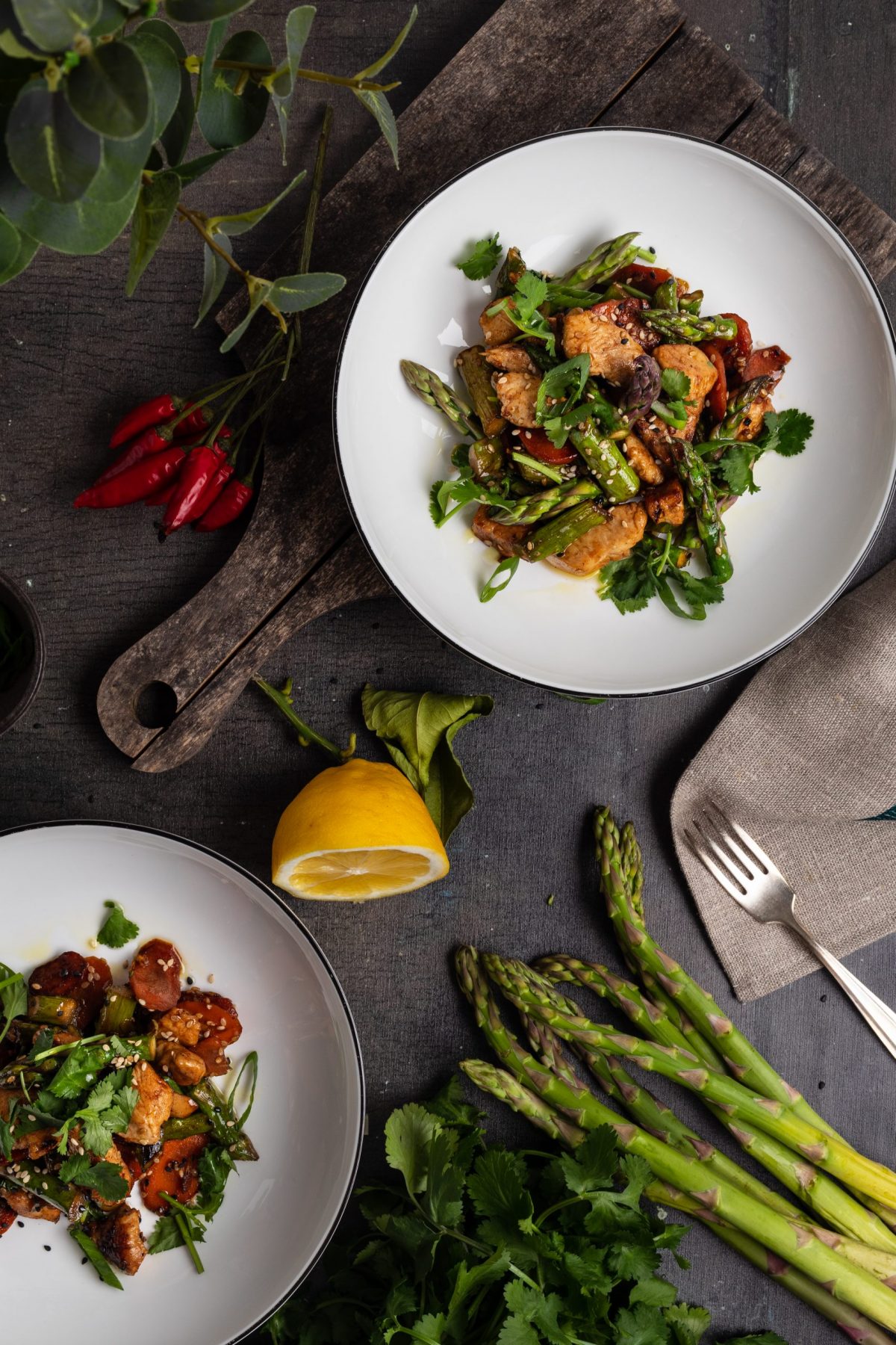 Zwei Teller mit Hühnchen, Spargel und Gemüse, umgeben von Zitronen, Chili und frischen Kräutern auf dunklem Holztisch.