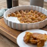 Ofenfrische Kartoffeln mit knuspriger Kruste in einer runden weißen Backform auf Holzbrett serviert.