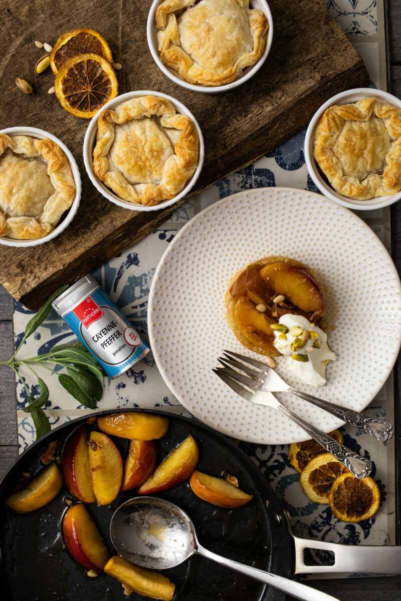 Verschiedene Dessert-Törtchen auf Holzbrett und Teller, dazu gebratene Apfelscheiben in Pfanne, dekoriert mit Gewürzen.