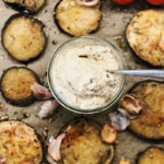 Ein Glas mit cremigem Aufstrich, umgeben von gebratenen Auberginenscheiben, Knoblauch und frischen Tomaten.
