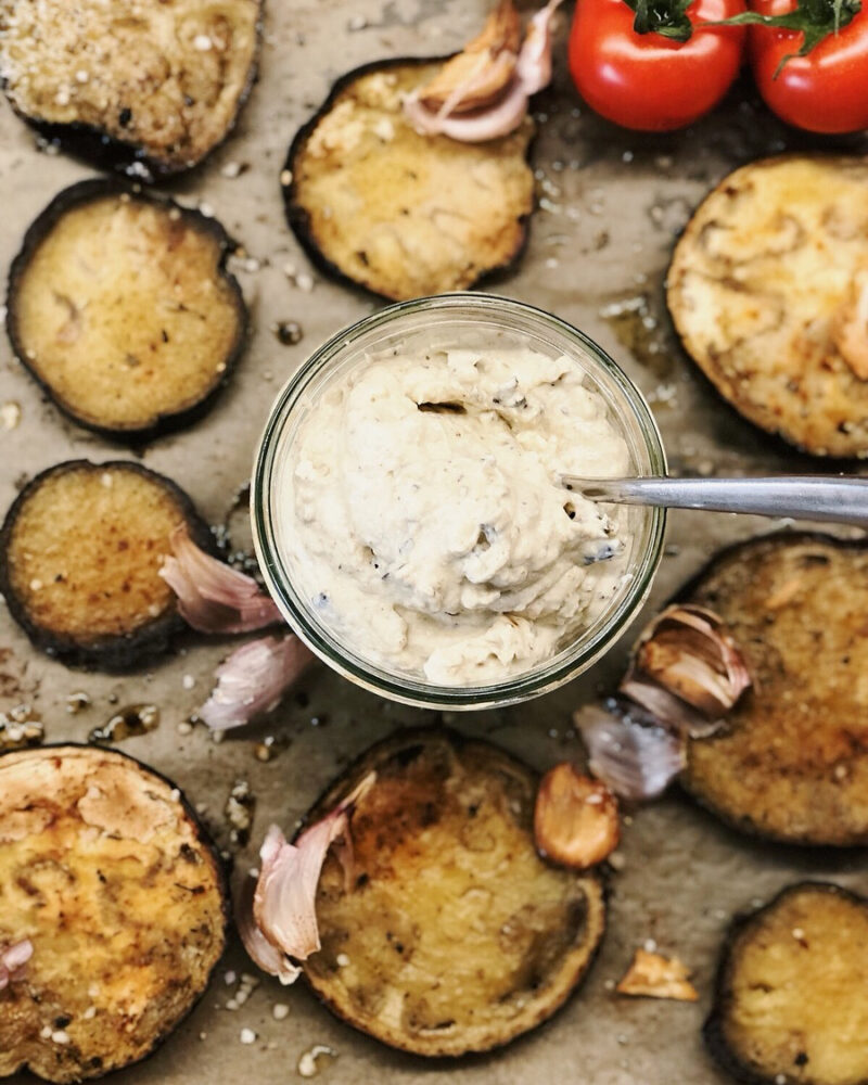 Ein Glas mit cremigem Aufstrich, umgeben von gebratenen Auberginenscheiben, Knoblauch und frischen Tomaten.