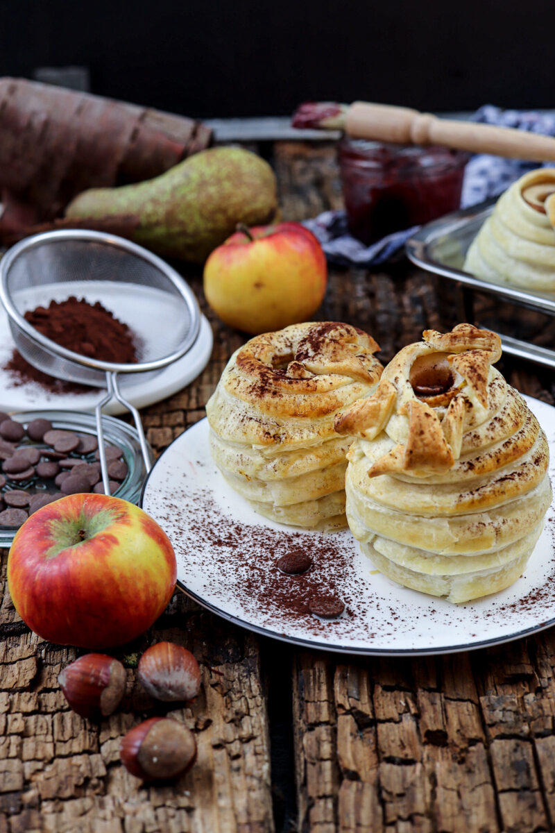 Zwei gebackene Apfel-Desserts auf einem Teller, bestäubt mit Kakao, umgeben von Äpfeln, Nüssen und Schokolade.
