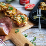 Holzbrett mit herzhaften Muffins aus Teig und Speck, Salami, Frühlingszwiebeln, Muffinform mit Gabel im Hintergrund.