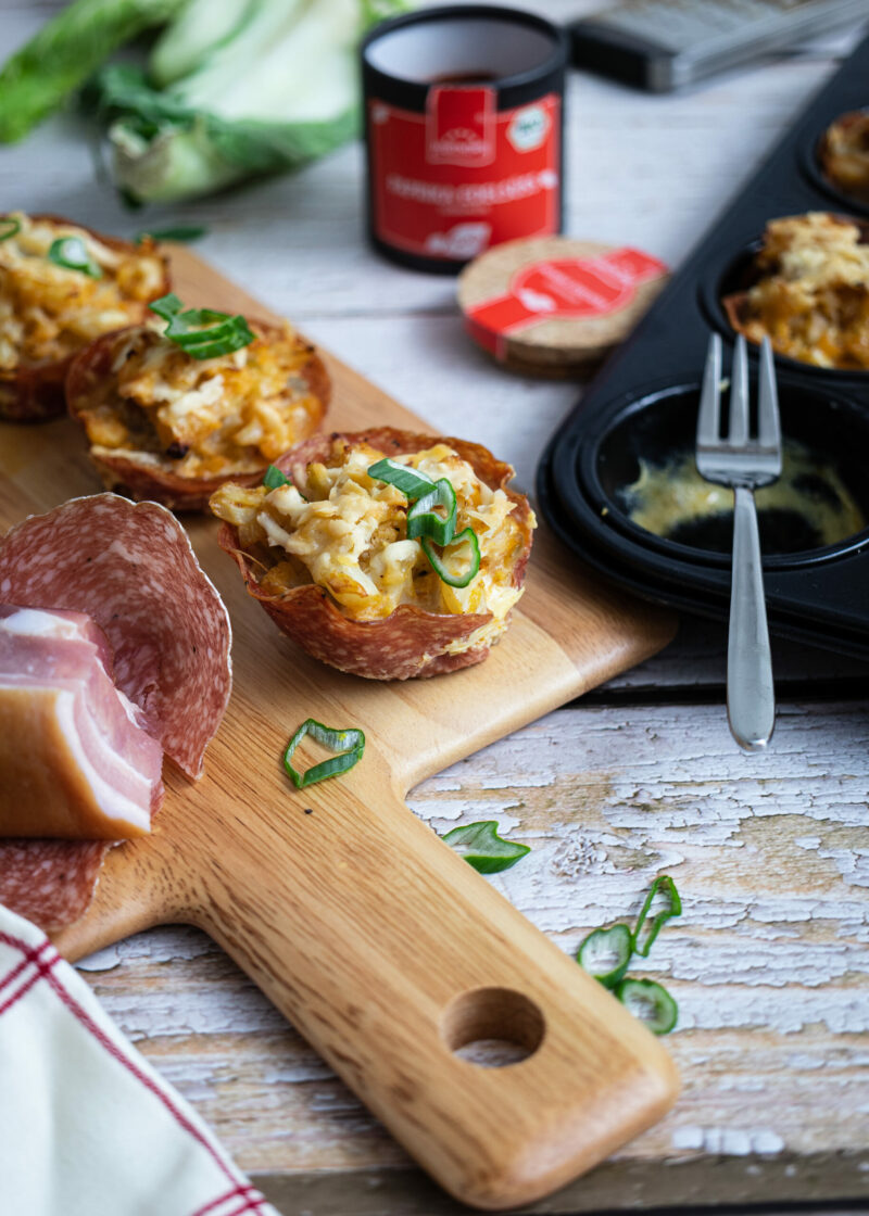 Holzbrett mit herzhaften Muffins aus Teig und Speck, Salami, Frühlingszwiebeln, Muffinform mit Gabel im Hintergrund.