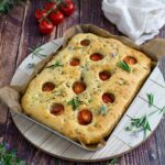 Focaccia mit Tomaten und Rosmarin auf Holzuntergrund, daneben frische Tomaten und ein weißes Tuch.