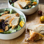 Zwei Schalen Caesar-Salat mit Hähnchen, Croutons, gelben Tomaten und Kräutern auf einem Holzbrett.