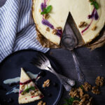 Ein angeschnittener Käsekuchen mit Beeren- und Nusssauce, daneben ein Stück auf einem Teller mit einer Gabel.