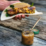 Hand hält Wrap mit Wurst, Petersilie und Cornflakes. Daneben Glas mit Sauce und Löffel auf Holztisch.