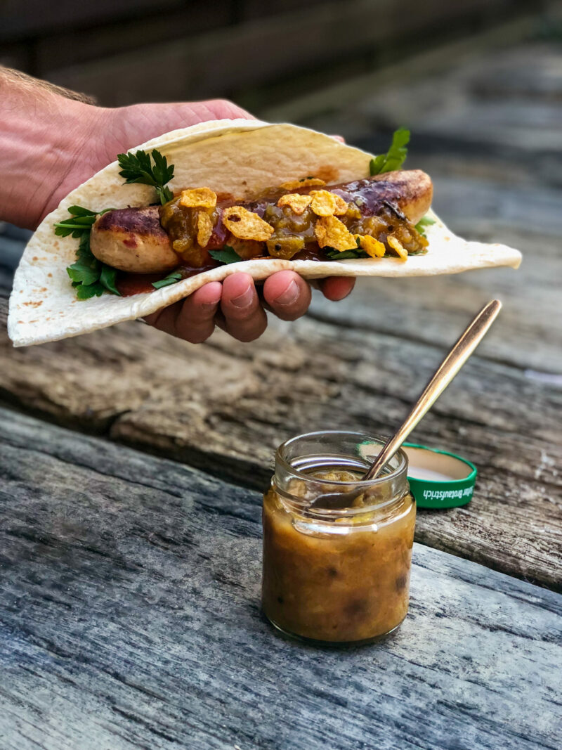 Hand hält Wrap mit Wurst, Petersilie und Cornflakes. Daneben Glas mit Sauce und Löffel auf Holztisch.