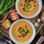 Zwei Schalen mit gelber Suppe, garniert mit Granatapfel und Frühlingszwiebeln. Gewürze, Besteck und Zutaten daneben.