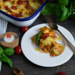 Überbackene, mit Käse und Tomaten belegte Pasta in einer Auflaufform, serviert auf einem Teller mit frischem Basilikum.