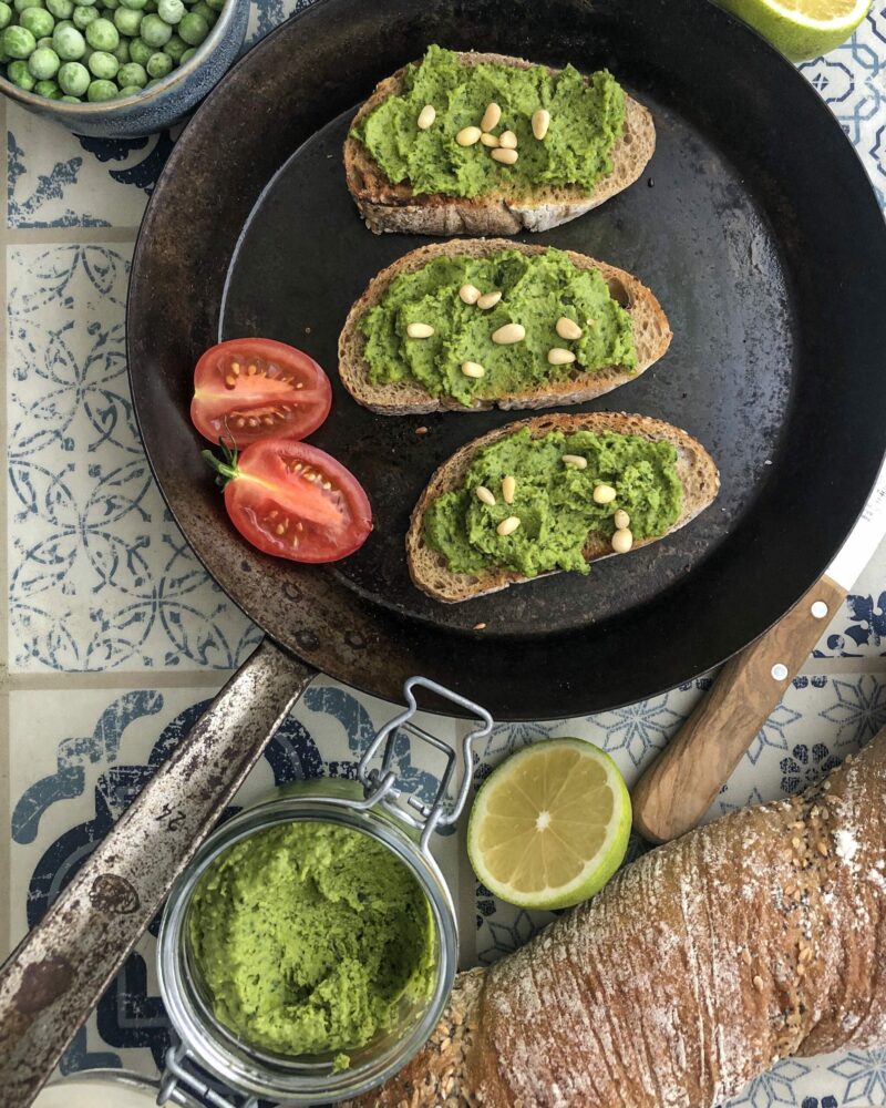 Drei Brotscheiben mit grünem Aufstrich und Pinienkernen in Pfanne, Tomatenhälfte, Glas, Zitrone und Baguette daneben.