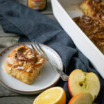 Zimtschnecke auf Teller neben Zitrone und Apfelscheiben, mit Backform und Gewürzdose im Hintergrund auf Holztisch.