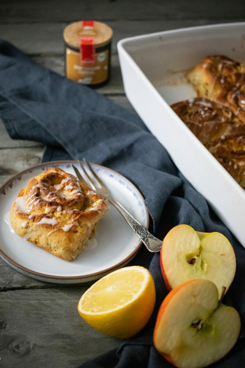 Zimtschnecke auf Teller neben Zitrone und Apfelscheiben, mit Backform und Gewürzdose im Hintergrund auf Holztisch.
