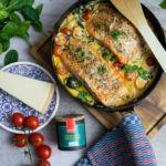 Gebackener Lachs mit Kirschtomaten und Spinat in Pfanne, umgeben von Käse und Gewürzen, auf weißem Hintergrund.