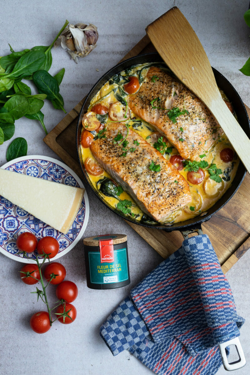 Gebackener Lachs mit Kirschtomaten und Spinat in Pfanne, umgeben von Käse und Gewürzen, auf weißem Hintergrund.