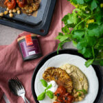Gerösteter Blumenkohl mit gebackenen Tomaten, Kichererbsen und Hummus auf einem Teller, daneben frische Kräuter und Gewürz.